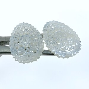 May include: Two white oval-shaped mother of pearl earrings with intricate floral carvings.