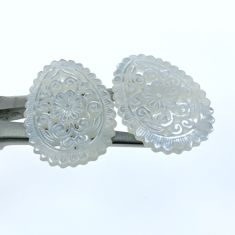 May include: Two white oval-shaped mother of pearl earrings with intricate floral carvings.