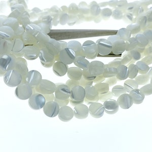 Może przedstawiać: Sznur białych, okrągłych koralików z muszli o perłowym, tęczowym połysku. Koraliki mają średnicę około 10 mm.