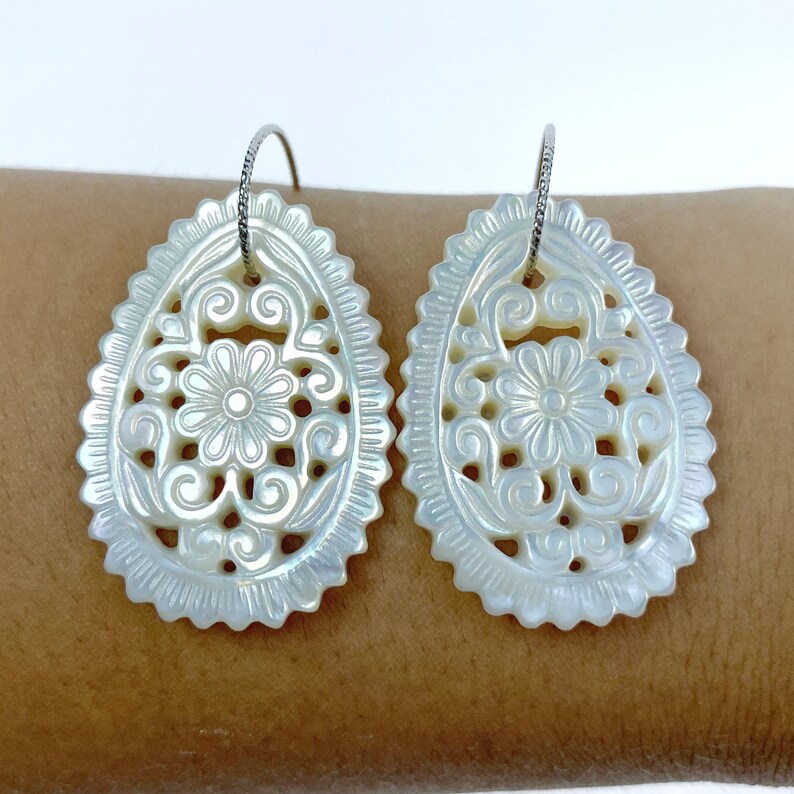 May include: A pair of white mother of pearl earrings with a floral design. The earrings are oval shaped and have a delicate, intricate pattern.