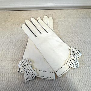May include: A pair of off-white gloves featuring black decorative stitching along the fingers and cuffs. Each glove is accented with a small, matching off-white bow at the wrist, which is also detailed with black dashed lines.