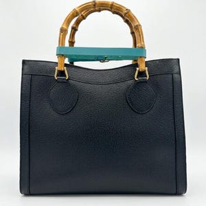 May include: A black leather handbag with a textured surface, featuring two rounded bamboo handles. A teal leather strap is wrapped around the base of the bamboo handles, secured with gold-toned metal hardware attachments on the top of the bag.