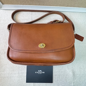 May include: A tan leather shoulder bag featuring a gold-tone turn-lock closure on the front flap and an adjustable strap. A small leather hangtag is attached to the side. A black card with the white text COACH NEW YORK is placed below.