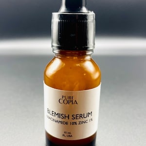 May include: A clear glass bottle with a black dropper top and a white label that reads "PURE COPIA BLEMISH SERUM NIACINAMIDE 10% ZINC 1%". The bottle contains a clear liquid and is labeled with "15 mL FL, USA".
