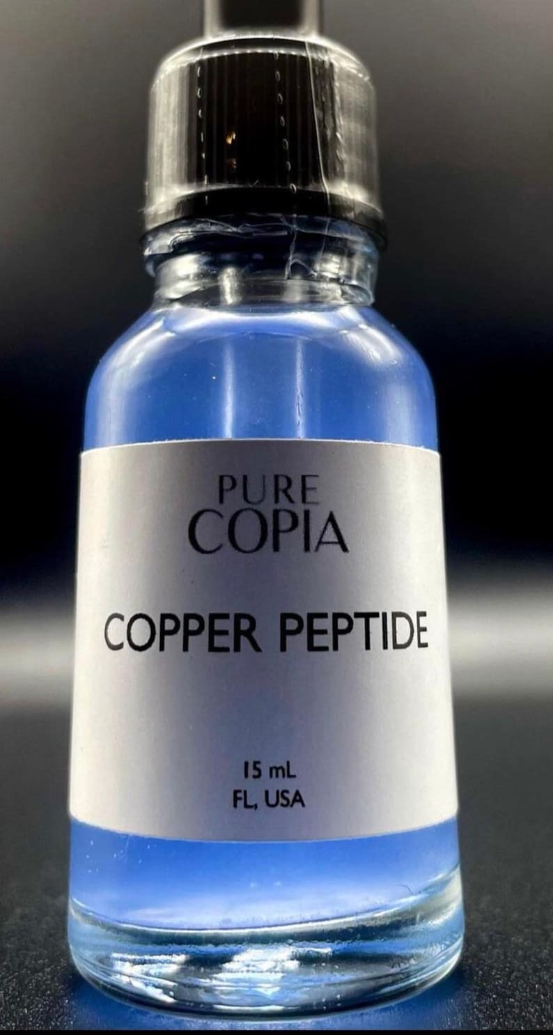 May include: A clear glass bottle with a blue liquid inside. The bottle has a white label with the text "PURE COPIA COPPER PEPTIDE" and "15 mL FL, USA".