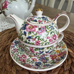 May include: A vintage tea set with a floral pattern. The set includes a teapot, teacup, and saucer, all decorated with pink, blue, and yellow flowers and green foliage on a white background. The teapot has a gold-colored handle and lid detail.