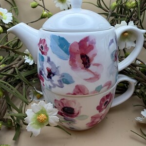 May include: A white ceramic teapot and teacup set with a floral pattern in pink, purple, and blue. The teapot has a lid and spout, and the teacup fits inside. Artificial flowers surround the set.