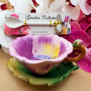 May include: A floral-themed teacup and saucer set with a matching spoon. The teacup is shaped like a purple and yellow flower, resting on a green leaf-shaped saucer. The spoon has a bee design and the text "Garden Collection".