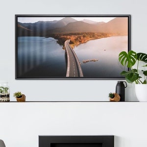 May include: A framed photograph of a winding road leading through a lake and mountains. The road is paved and the lake is surrounded by trees. The mountains are in the background and the sky is a light blue.