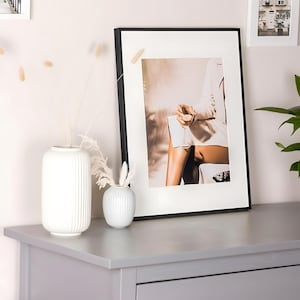 Könnte beinhalten: Ein schwarzer gerahmter Druck einer Frau, die auf einem Stuhl sitzt und ein weißes Kleid trägt. Der Hintergrund ist ein weiches, verschwommenes Bild eines Raumes mit einfallendem Licht. Der Druck befindet sich auf einer grauen Kommode mit einer weißen Vase und einer kleinen weißen Schale.