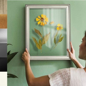 Könnte beinhalten: Eine Frau hängt ein gerahmtes Bild mit gepressten Blumen an einer grünen Wand. Der Rahmen ist silberfarben mit einem weißen Passepartout und die Blumen sind gelb und braun.