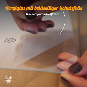 May include: A hand peeling off a clear protective film from a sheet of acrylic glass. The text on the film reads "Acrylglas mit beidseitiger Schutzfolie Bitte vor Gebrauch entfernen".