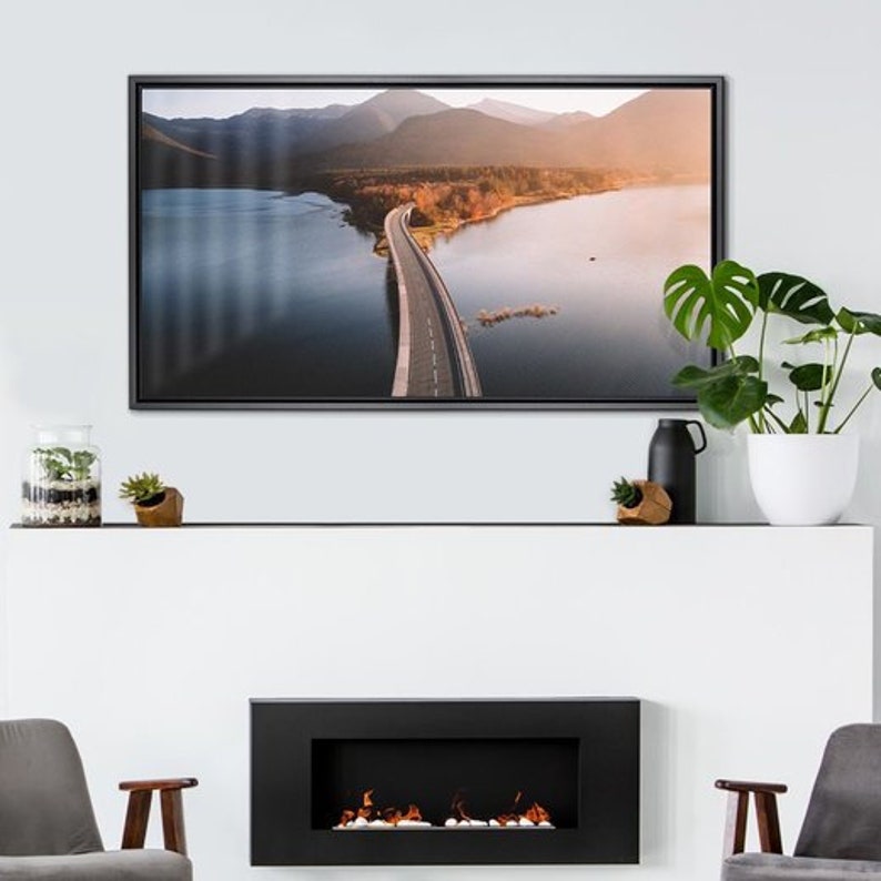May include: A framed photograph of a winding road leading through a lake and mountains. The road is paved and the lake is surrounded by trees. The mountains are in the background and the sky is a light blue.