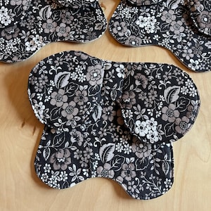 May include: Several reusable cloth menstrual pads with a floral pattern in black, white, and taupe. The pads are in various shapes and sizes, with rounded and curved edges. The fabric has a dense floral print, and the pads are arranged on a light wood surface.