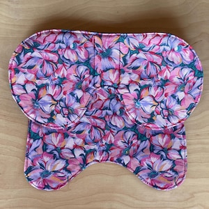 May include: A floral-patterned saddle pad featuring a vibrant design of pink, purple, and teal flowers. The pad has a contoured shape with rounded edges and a central cutout, likely for the horse's spine. The fabric appears to be a soft, quilted material.