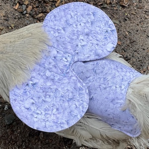 May include: A light gray chicken wearing a light purple chicken saddle. The saddle is made of fabric with a floral pattern. The chicken is on a brown surface.