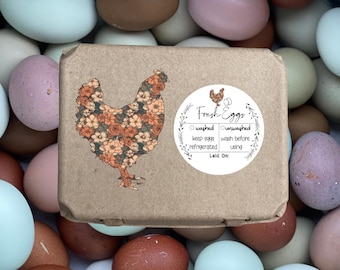 May include: A brown cardboard box with a floral patterned chicken illustration and a sticker that reads "Fresh Eggs - Washed - Unwashed - Keep eggs refrigerated - Wash before using - Laid On". The box is surrounded by a variety of brown, blue, and pink eggs.