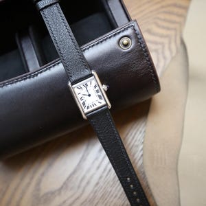 Könnte beinhalten: Eine silberne rechteckige Armbanduhr mit weißem Zifferblatt und schwarzen römischen Ziffern. Die Uhr hat ein schwarzes Lederarmband. Die Uhr befindet sich neben einem dunkelbraunen Leder-Uhrenetui.
