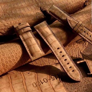 Könnte beinhalten: Hellbraune Leder-Armbänder mit Krokodilhautmuster. Die Armbänder haben sichtbare Nähte und eine Schnalle. Der Hintergrund zeigt mehr Leder mit der gleichen Textur. Die Armbänder sind neu.