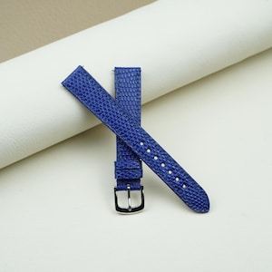 Può includere: Un cinturino per orologio blu vivace caratterizzato da una trama in rilievo simile alla pelle di lucertola. Il cinturino include una fibbia in metallo argentato lucido e un passante blu coordinato, con fori per la regolazione.