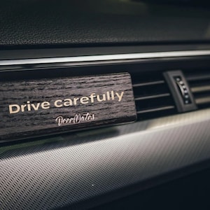 Puede incluir: Un ambientador de coche de madera con el texto "Conduce con cuidado" y el nombre de la marca "WoodNotes" en una fuente dorada.