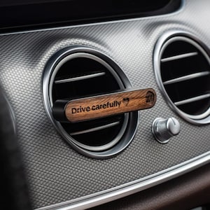 Könnte beinhalten: Ein hölzerner Autoluftaufbereiter mit dem Text "Fahren Sie vorsichtig" und einem Herzsymbol. Der Luftaufbereiter ist an einer Lüftung eines Autos befestigt.