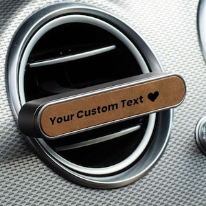 Peut inclure: Un support de téléphone pour grille d'aération de voiture avec une surface en similicuir marron et le texte "Your Custom Text" avec un symbole de cœur. Le support est noir et argenté.