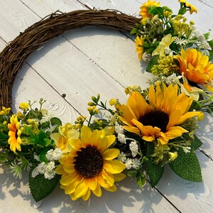 May include: A brown twig wreath decorated with yellow sunflowers and white flowers. The wreath is on a white wooden background.
