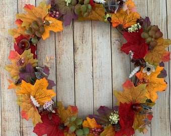 May include: A fall-themed wreath made of artificial leaves in shades of red, orange, yellow, and green. The wreath is decorated with small white flowers and berries.