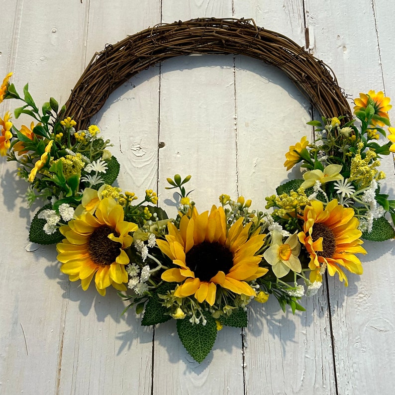 May include: A sunflower wreath with a brown vine base. The wreath is decorated with yellow sunflowers, white flowers, and green leaves.