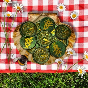 Puede incluir: Una colección de monedas decorativas verdes y doradas con varios diseños, exhibidas en una gran concha. Las monedas están dispuestas sobre una manta de picnic a cuadros rojos y blancos, rodeadas de margaritas y un abejorro.