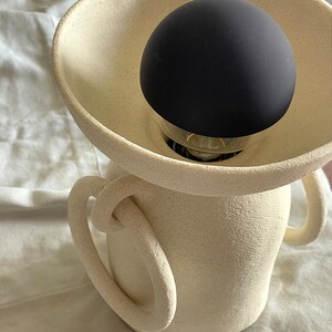 Pu&ograve; includere: Una lampada in ceramica bianca con due maniglie e un paralume nero a forma di globo.