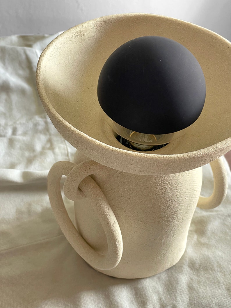 Pu&ograve; includere: Una lampada in ceramica bianca con due maniglie e un paralume nero a forma di globo.
