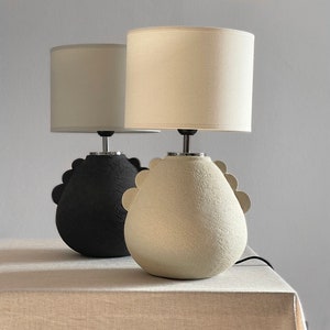 table lamp, the lower part is a hemisphere, the upper part is conical where the lamp holder and the lamp shade are located, the base has on both sides semi-disc shaped ornaments, it looks like a geometric hen's crepe. Porous texture.