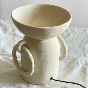 Pu&ograve; includere: Una lampada in ceramica bianca con una parte superiore a forma di ciotola e due manici. La lampada ha una superficie testurizzata e un cavo nero.