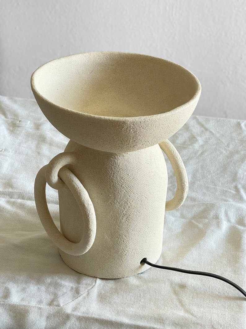 Pu&ograve; includere: Una lampada in ceramica bianca con una parte superiore a forma di ciotola e due manici. La lampada ha una superficie testurizzata e un cavo nero.