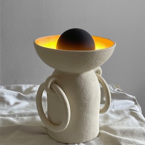 Pu&ograve; includere: Una lampada in ceramica bianca con due manici e una parte superiore a forma di ciotola. La ciotola ha una luce incandescente all'interno e una sfera nera al centro.
