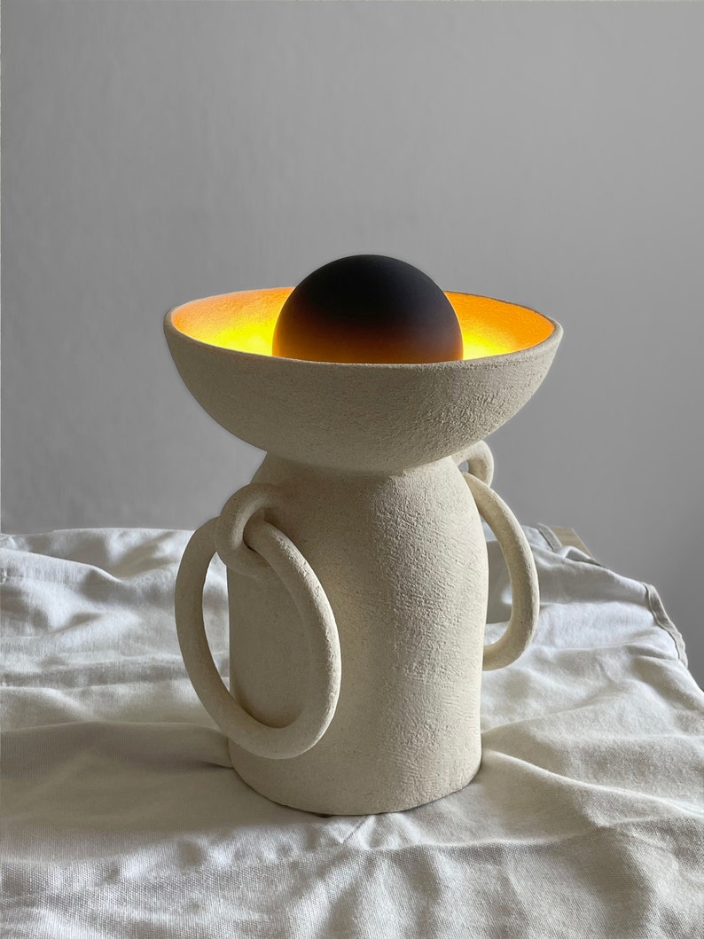 Pu&ograve; includere: Una lampada in ceramica bianca con due manici e una parte superiore a forma di ciotola. La ciotola ha una luce incandescente all'interno e una sfera nera al centro.