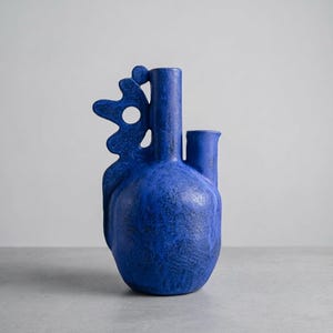 May include: A vibrant blue ceramic vase with a textured, matte finish. The vessel features a rounded base, a tall central neck, and a shorter secondary spout. An abstract, organic-shaped handle with a circular cutout extends from the side.