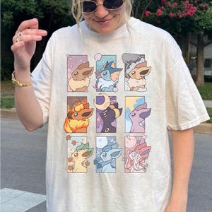 May include: A cream-coloured short-sleeve t-shirt featuring a grid of nine rectangular panels, each displaying a different stylised cartoon creature character in various pastel-coloured settings, including floral, aquatic, stormy, fiery, and lunar themes.