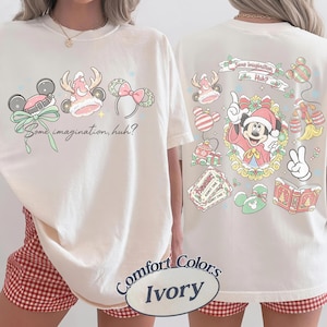 以下が含まれることがあります： アイボリー色のTシャツ。前面にはホリデーをテーマにした3つのマウスイヤーカチューシャと「Some imagination, huh?」の文字がプリントされています。背面にはサンタ帽をかぶったキャラクターを中心に、お祝いのアイコンが描かれています。