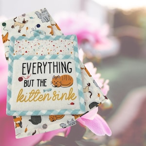 May include: A set of two blue and white kitchen towels with a cat-themed design. The towels feature a white background with a blue and white checkered border. The front of the towels features a cat design with the text "Everything but the kitten sink".