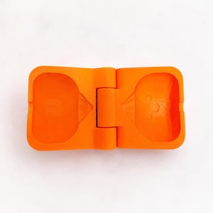 May include: A bright orange, rectangular, hinged plastic mold featuring two symmetrical, hollowed-out cavities. Each cavity has a distinct, recessed shape with a small, circular indentation at the bottom, designed for creating molded items.