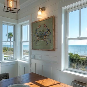 May include: A square framed wall art piece featuring a colorful, stylized illustration of a blue crab centered on a muted olive green background, mounted on a white shiplap wall between two windows overlooking a coastal beach scene.