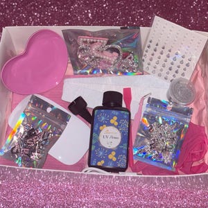 May include: A nail art kit in a white box, containing a pink heart-shaped dish, nail gems, a UV gel bottle, and manicure tools. The kit includes a nail file, glitter, and a pink spatula. The background is a sparkly pink surface.