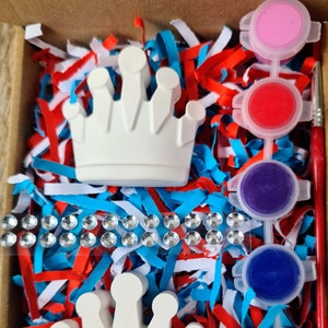 May include: Two white ceramic crowns, one with a black circle in the centre, in a box with red, white and blue shredded paper. There are eight small paint pots with different colours of paint.