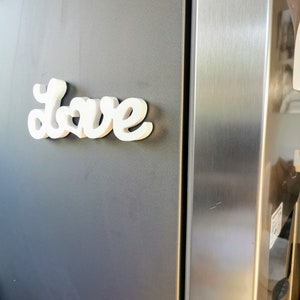 May include: White, three-dimensional letters spelling "Love" are attached to a grey refrigerator door.