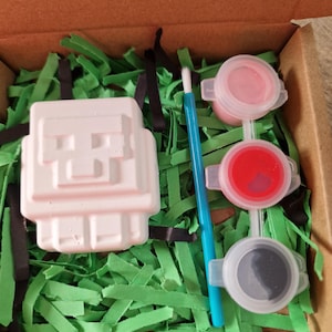 May include: A craft kit in a cardboard box, featuring a white, blocky figure, three small paint pots with pink, red, and gray paint, a blue paintbrush, and green shredded paper. The kit is designed for creative activities.