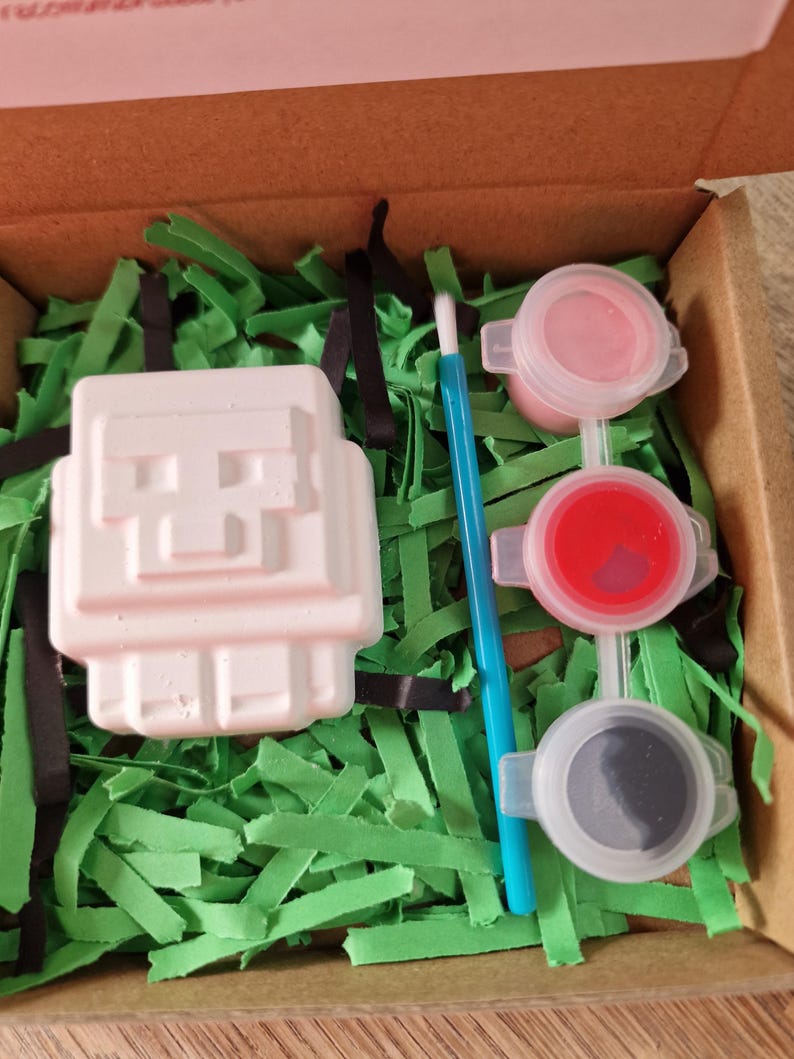 May include: A craft kit in a cardboard box, featuring a white, blocky figure, three small paint pots with pink, red, and gray paint, a blue paintbrush, and green shredded paper. The kit is designed for creative activities.