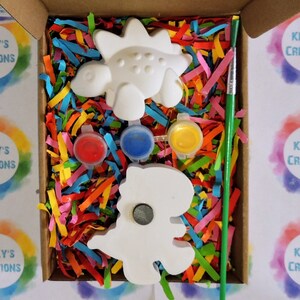 May include: A craft kit with two white ceramic dinosaur figurines, paint pots in red, blue, and yellow, and a green paintbrush. The kit is packaged in a cardboard box with colorful shredded paper.